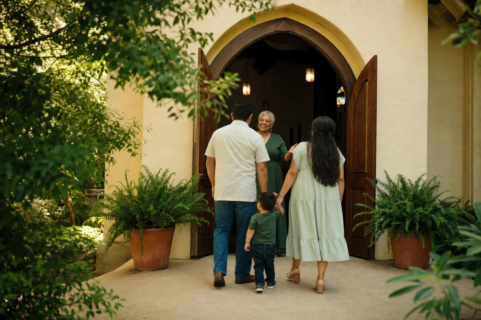 A silver-haired greeter welcoming a young family at the chapel doorway framed by potted ferns